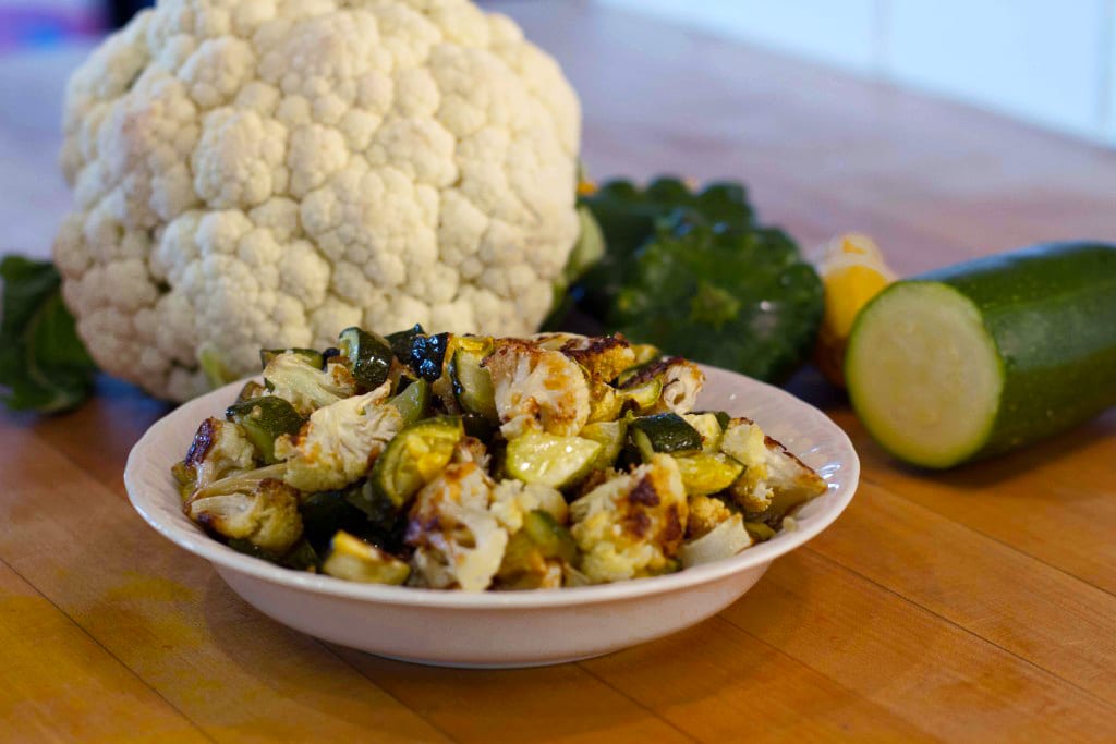 fresh cauliflower sits on table next to freshly cut cucumber and bowl of garlic roasted cauliflower